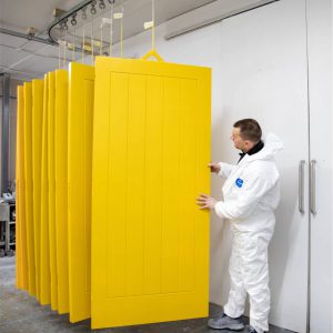 Man in white overalls stood in a spray painting workshop in front of several yellow, hanging doors